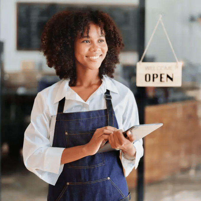 Young woman with curly dark hair holding a clipboard next to an open sign, with a cafe in the background